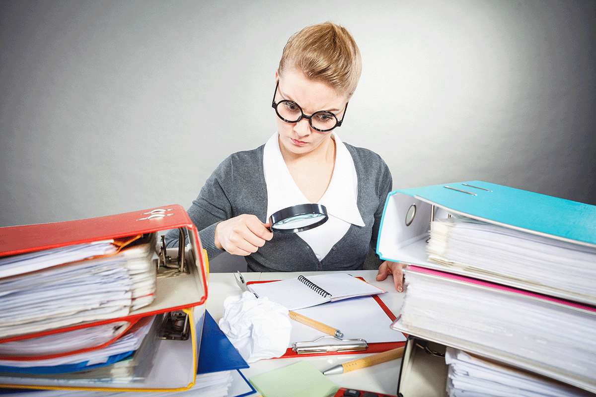 Young lady holding a magnifying glass looking at an open notebook on a desk full of binders