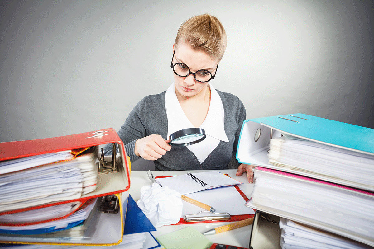 A young lady in round glasses holding a magnifying glass looking at an open notebook that is on the desk full of binders.