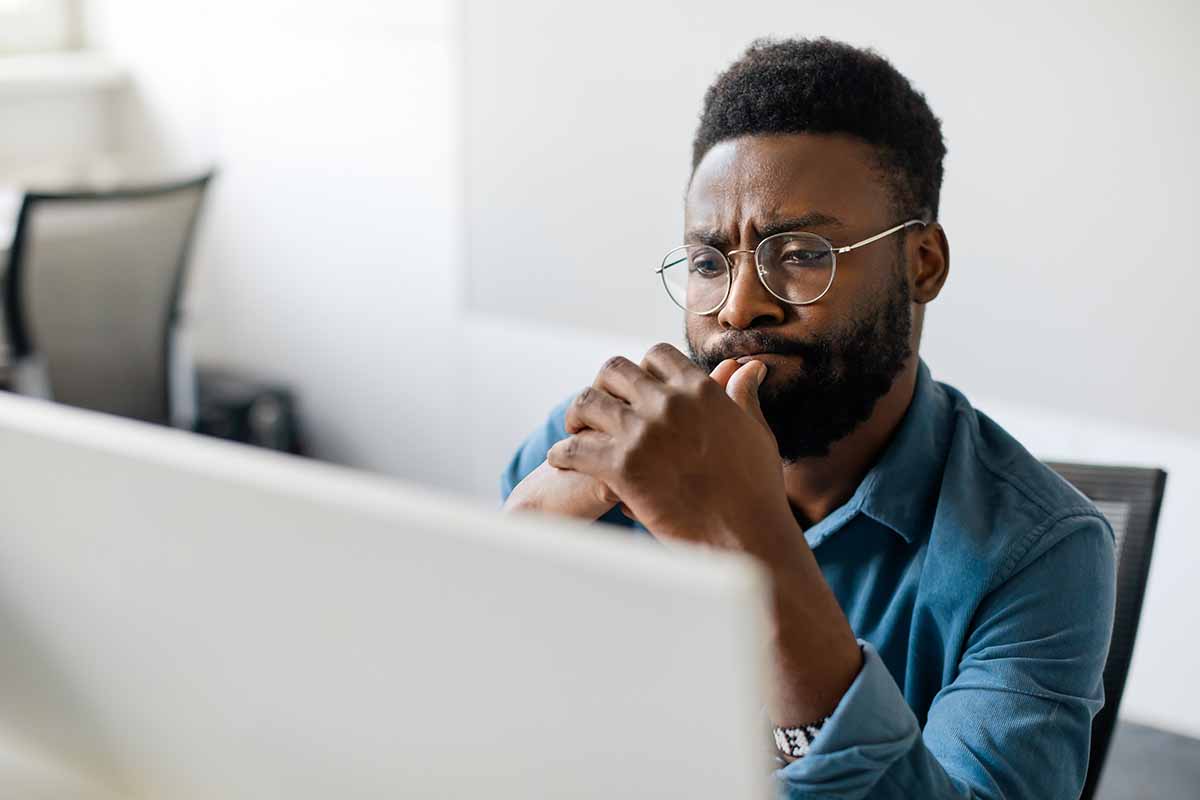 A young African American man in eyeglasses deep in thought looking at computer screen.