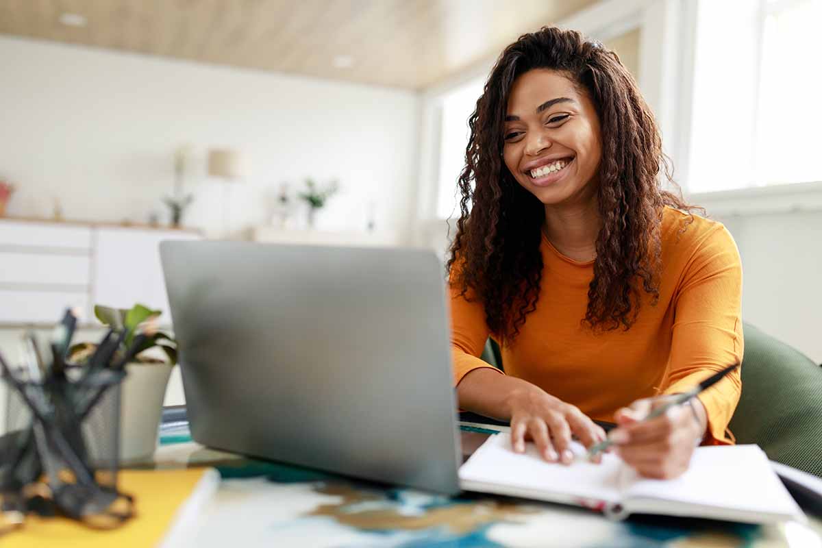 An African American smiling woman looking at a laptop while taking notes.