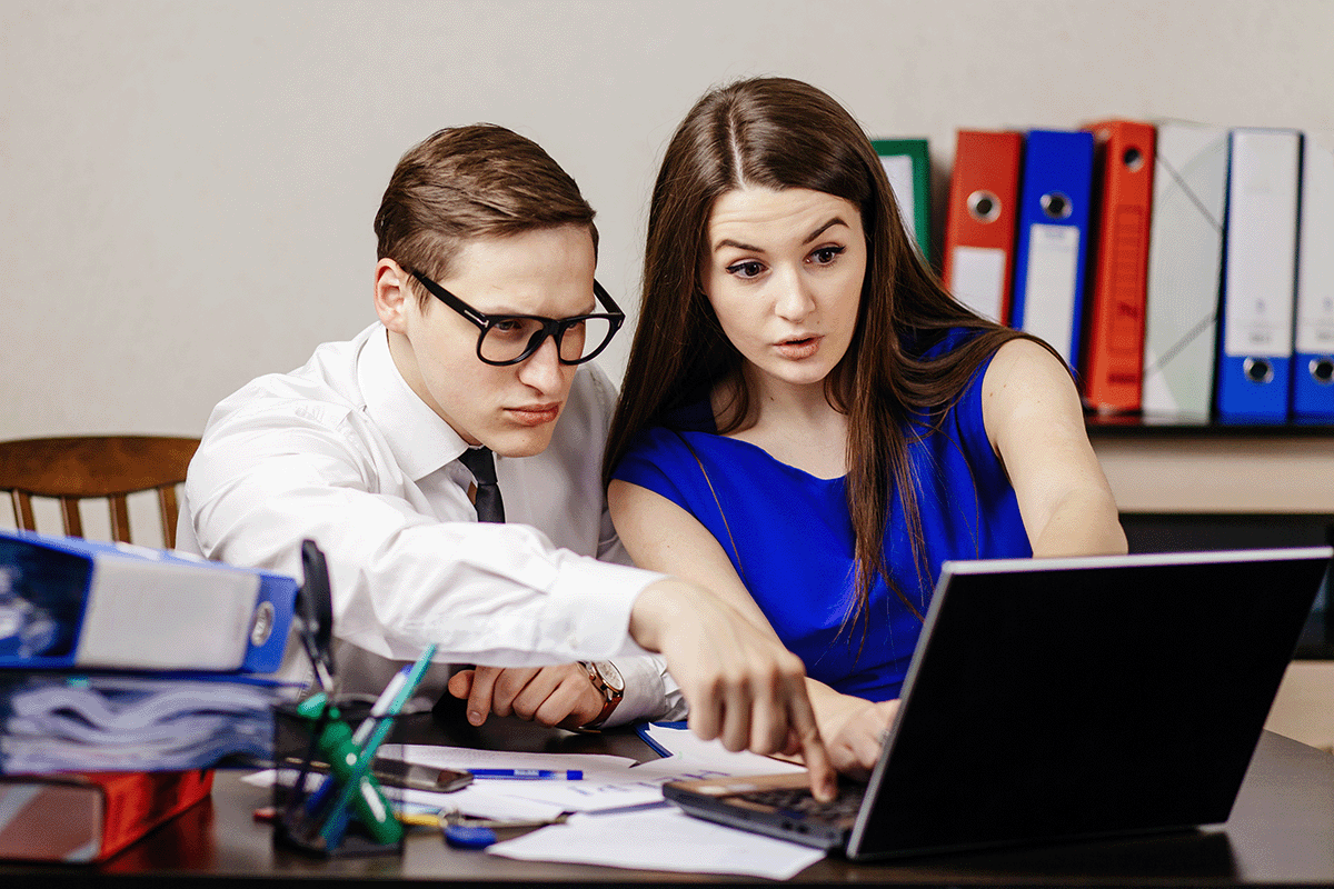 Two young business coworkers with a puzzled look on their faces sitting at a desk looking at an open laptop screen.