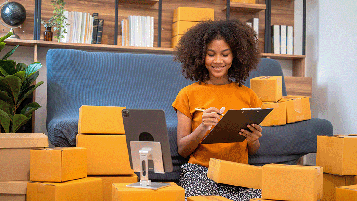 An African American business woman with boxes surrounding here, while she is holding a clipboard and pen.