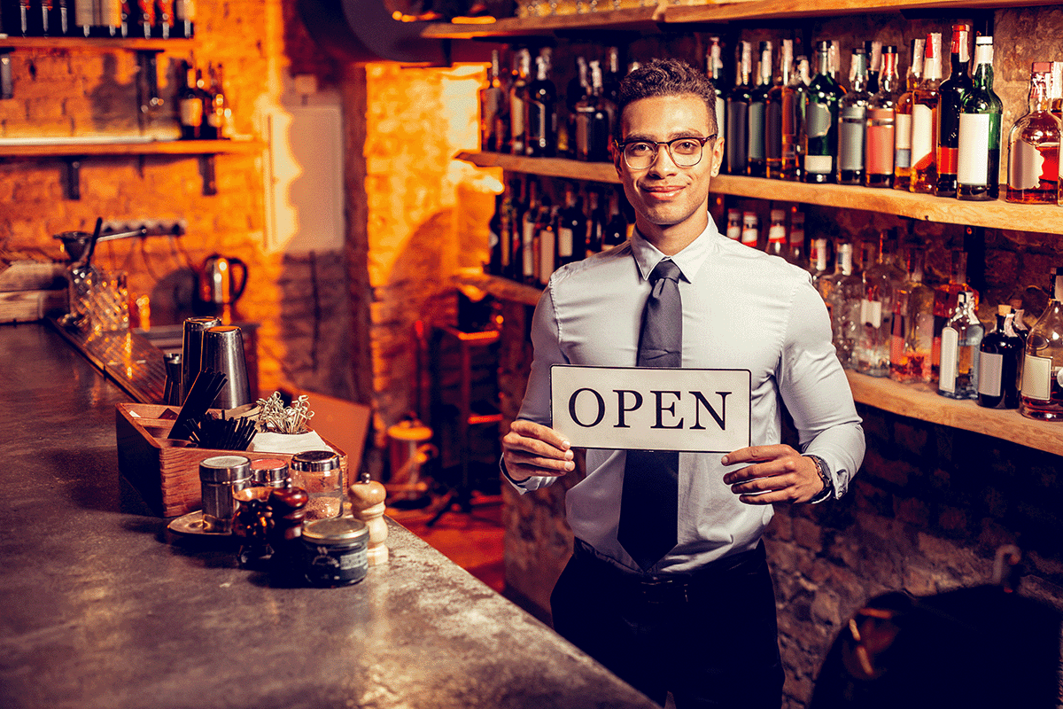 A smiling young African American businessman dressed in a dress shirt and tie behind a bar holding an open sign.