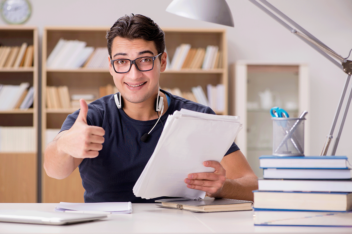 A young man smiling and giving a thumbs up, holding some paperwork sitting at a desk that is full of books, a pen holder, and a desk lamp.